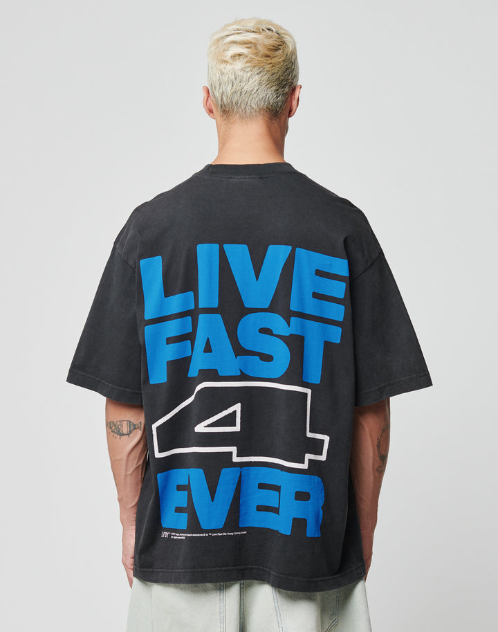A person with short, bleached hair wears the LIVE FAST DIE YOUNG LFDY 4 Ever Tee, an oversize black Baumwolle T-shirt with a bold blue and white LIVE FAST 4 EVER backprint, as they face away from the camera. | model