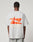 A person wears the LIVE FAST DIE YOUNG LFDY Hardwear Tee, a light gray oversized fit t-shirt featuring an orange abstract graphic and LIVE FAST™ HARDWEAR on the back, standing before a plain light background.