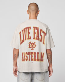 A person with a shaved head, seen from behind, wears the beige Live Fast Amsterdam Tee by LIVE FAST DIE YOUNG, featuring bold red and yellow LIVE FAST and AMSTERDAM text with a circular logo in between.