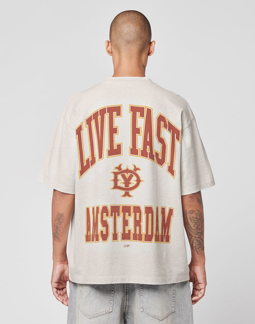 A person with a shaved head, seen from behind, wears the beige Live Fast Amsterdam Tee by LIVE FAST DIE YOUNG, featuring bold red and yellow LIVE FAST and AMSTERDAM text with a circular logo in between.