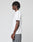 A young person with short twists stands in profile against a plain background, wearing the LIVE FAST DIE YOUNG Live Fast Plain Tee, a white frontprint t-shirt made from 100% Baumwolle, paired with gray pants.