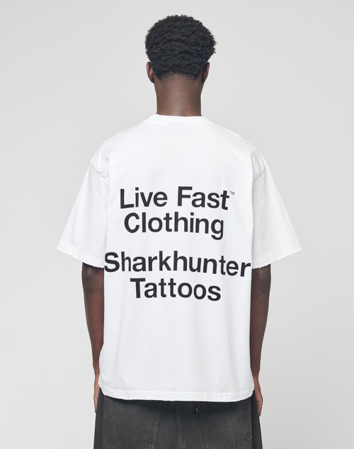 A person stands facing away, wearing the LFDY Sharkhunter Tee by LIVE FAST DIE YOUNG—an oversize white shirt with black Sharkhunter Tattoos text. Made from 100% Baumwolle, it’s set against a plain light gray background.