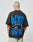 A person with short, bleached blond hair faces away wearing the LIVE FAST DIE YOUNG LFDY 4 Ever Tee, an oversized black shirt with bold blue and white LIVE FAST 4 EVER backprint, paired with light shorts and revealing a tattooed arm.