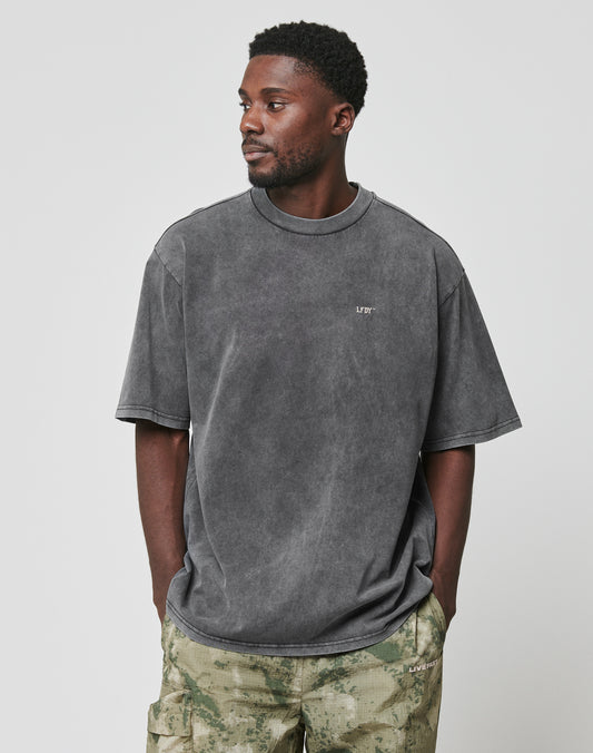 A man wears the LIVE FAST DIE YOUNG LFDY Property Tee, an oversized faded gray 100% Baumwolle shirt with small white chest text, paired with green camo pants. He stands against a plain light gray background, looking left with hands in pockets.