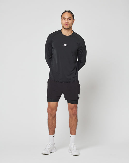 A person stands against a plain white background, wearing LIVE FAST DIE YOUNGs black Live Fast Performance Running Shorts with integrated tights, a black long-sleeve athletic shirt, white socks, and white sneakers. Hands behind back, facing forward.