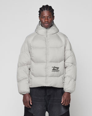A man with braided hair wears the LIVE FAST DIE YOUNG Live Fast Puffer Jacket, a light gray, oversized, hooded style featuring a reflektierender Print and LIVE FAST graphic, standing against a plain white background.