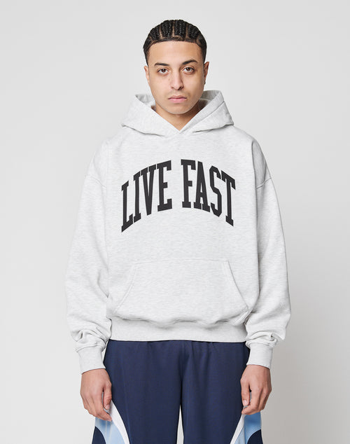 A person with braided hair wears a LIVE FAST DIE YOUNG Live Fast College Hooded in oversize light gray, featuring bold black LIVE FAST text, paired with dark blue athletic pants, standing against a plain white background.