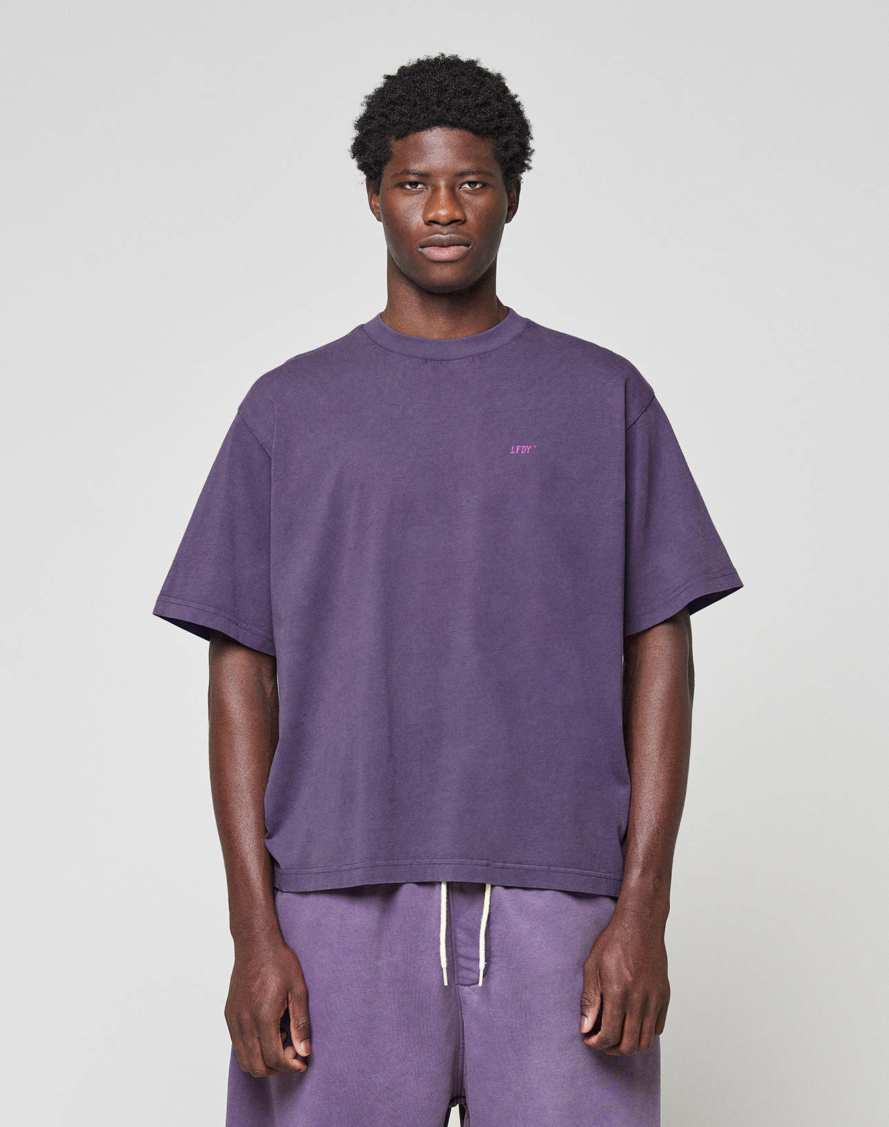 A man stands against a plain light background wearing the LIVE FAST DIE YOUNG Basic Evidence Tee in purple and matching drawstring shorts made from 100% Baumwolle, looking straight at the camera with a neutral expression.