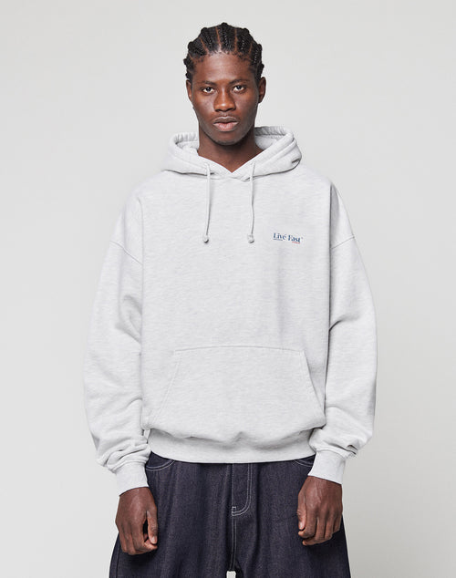 A person with braided hair wears a light gray, oversized fit Live Fast Paris Hooded by LIVE FAST DIE YOUNG, featuring a small text logo on the left chest. They stand against a plain light gray background with a neutral expression.