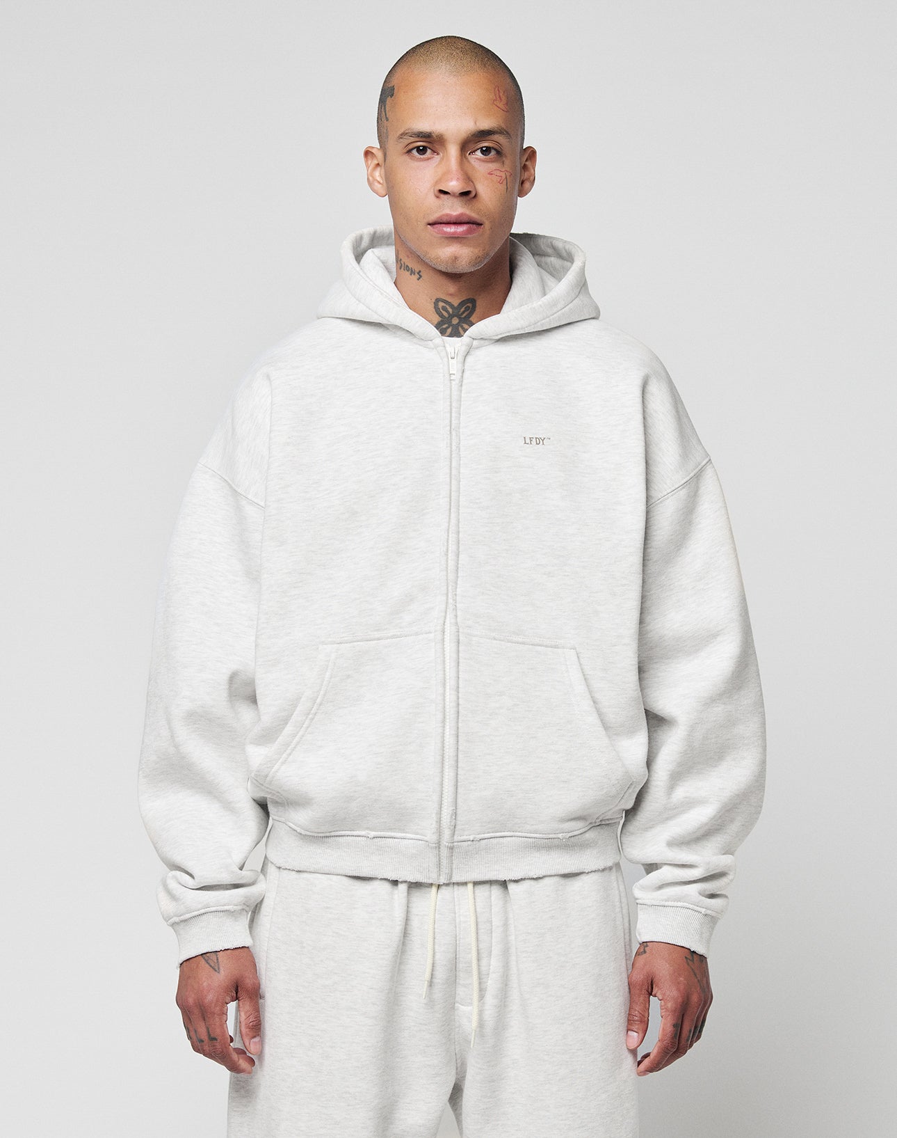 A person with a shaved head and visible tattoos wears the LIVE FAST DIE YOUNG Basic Hooded Zip in light gray with matching sweatpants, standing against a plain light gray background.