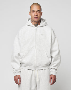 A person with a shaved head and visible tattoos wears the LIVE FAST DIE YOUNG Basic Hooded Zip in light gray with matching sweatpants, standing against a plain light gray background.