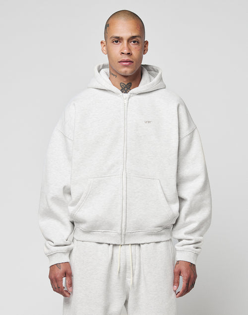 A person with a shaved head and visible tattoos wears the LIVE FAST DIE YOUNG Basic Hooded Zip in light gray with matching sweatpants, standing against a plain light gray background.