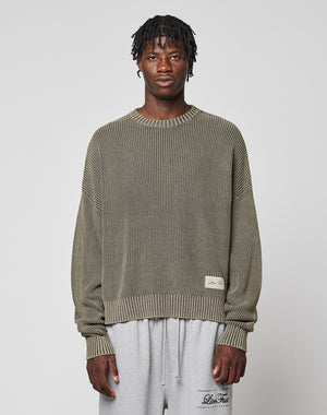 A man with short twists wears the LIVE FAST DIE YOUNG LFDY Knit Sweater, an oversized ribbed olive-green piece in 100% cotton, paired with light gray sweatpants. He stands facing forward against a plain light gray background.