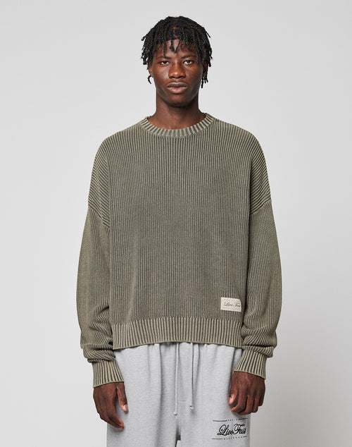 A man with short twists wears the LIVE FAST DIE YOUNG LFDY Knit Sweater, an oversized ribbed olive-green piece in 100% cotton, paired with light gray sweatpants. He stands facing forward against a plain light gray background.