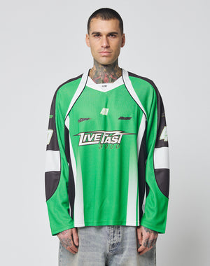 A person with short hair and visible tattoos wears the LIVE FAST DIE YOUNG LFDY Motorcross Jersey—an oversized green, black, and white top with multi-logo prints, Live Fast 3000, and the number 40—against a plain light background.