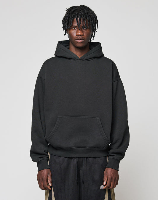 A person with short, twisted hair faces forward, wearing a black Basic Blank Hooded by LIVE FAST DIE YOUNG and black shorts with beige stripes and a flag label, set against a plain light gray background.