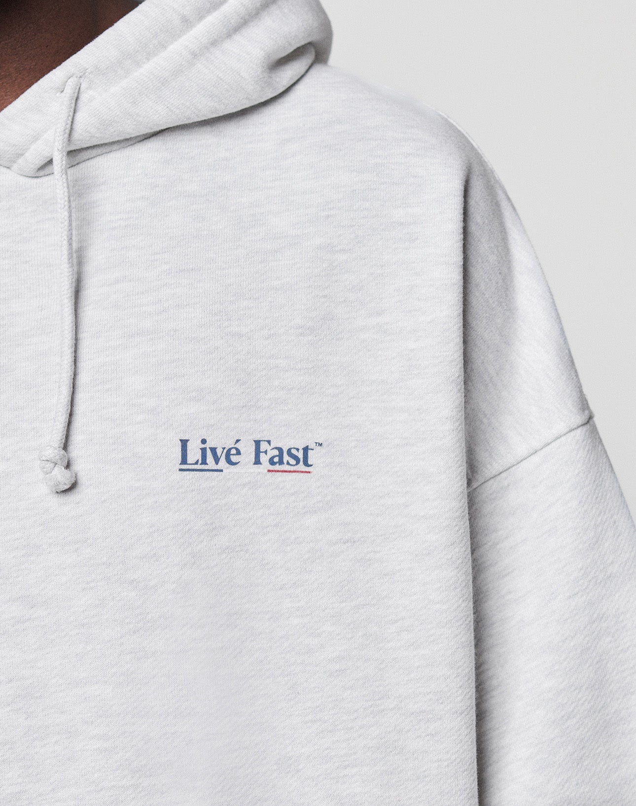 Close-up of the Live Fast Paris Hooded by LIVE FAST DIE YOUNG, a light gray cotton hoodie with an oversized fit. Features "Live Fast" in small blue and pink letters on the left chest, drawstring hood, visible stitching, and ultra-comfy style.
