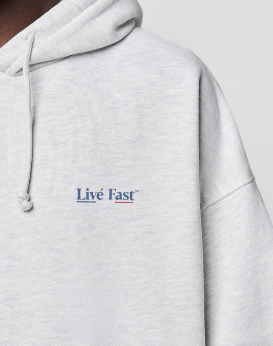 Close-up of the Live Fast Paris Hooded by LIVE FAST DIE YOUNG, a light gray cotton hoodie with an oversized fit. Features "Live Fast" in small blue and pink letters on the left chest, drawstring hood, visible stitching, and ultra-comfy style.
