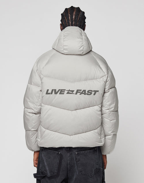 A person with braided hair models the LIVE FAST DIE YOUNG Live Fast Puffer Jacket in light grey. The oversize, wasserabweisend jacket features “LIVE FAST” in bold, reflektierender Print across the back. Seen from behind on a plain white background.