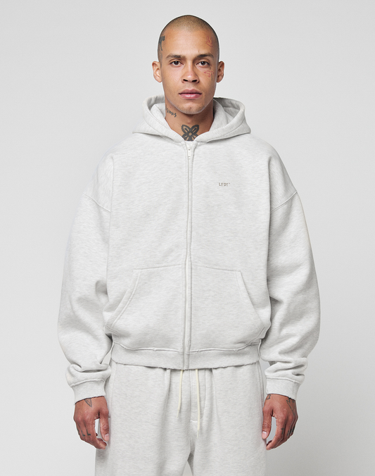 A person with a shaved head and visible tattoos models the LIVE FAST DIE YOUNG Basic Hooded Zip in light gray, paired with matching sweatpants, standing against a plain background and looking directly at the camera with a neutral expression.