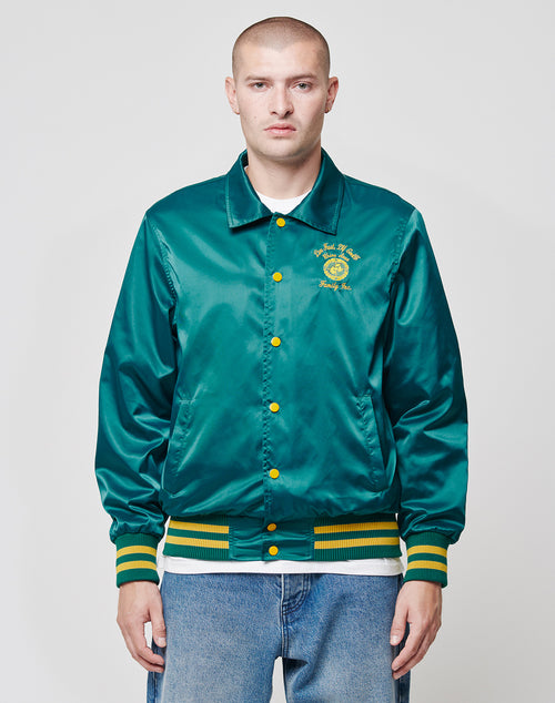 A man with a shaved head wears the LIVE FAST DIE YOUNG Rhine Area College Jacket—a shiny green varsity style with yellow buttons and striped cuffs—over a white shirt and regular fit blue jeans, standing against a plain background with a neutral expression.