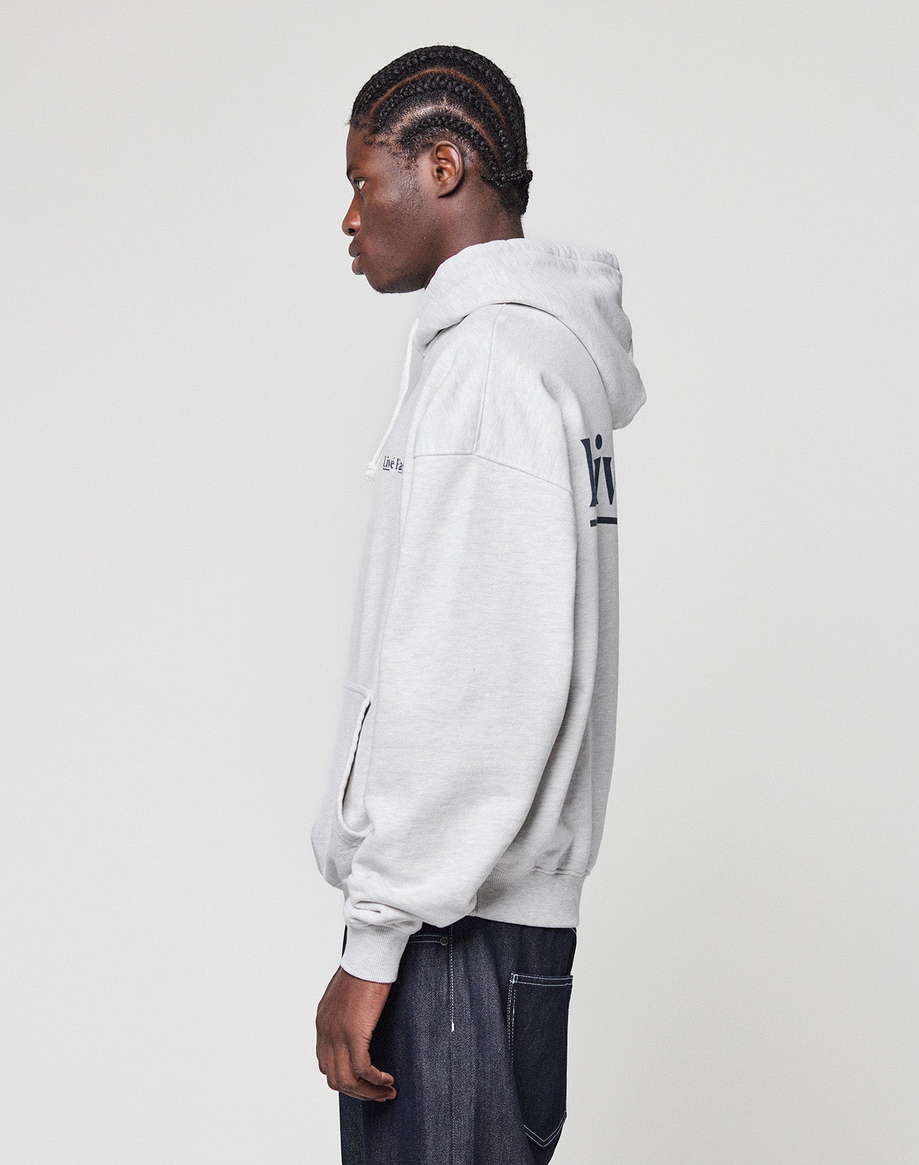 A person with braided hair stands in profile, wearing a light gray Live Fast Paris Hooded pullover by LIVE FAST DIE YOUNG and dark pants, set against a plain, light background.