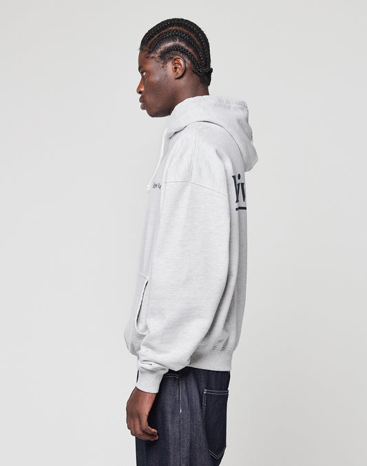 A person with braided hair stands in profile, wearing a light gray Live Fast Paris Hooded pullover by LIVE FAST DIE YOUNG and dark pants, set against a plain, light background.