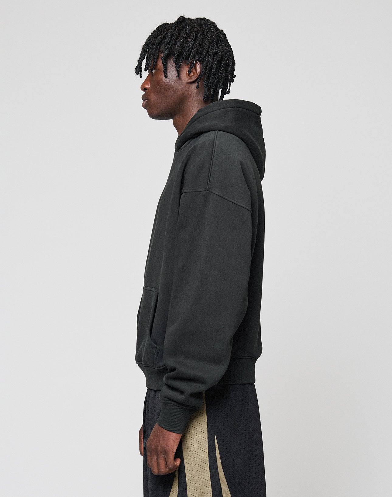 A person with short, twisted hair wears the LIVE FAST DIE YOUNG Basic Blank Hooded in an oversize fit, paired with black pants featuring a tan stripe, standing in profile against a plain light gray background.