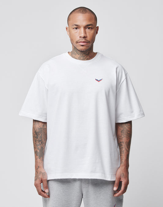 A man with tattooed arms faces forward, wearing the LFDY x Trigema Tennis Tee by LIVE FAST DIE YOUNG—an oversized white T-shirt with a small colorful embroidered chest logo—and light gray sweatpants, set against a plain light gray background.