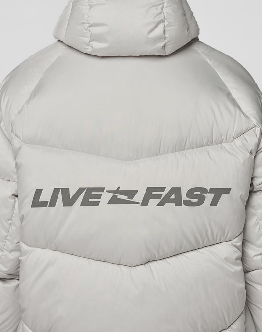 A close-up of the Live Fast Puffer Jacket by LIVE FAST DIE YOUNG shows the light gray, oversized fit with “LIVE FAST” in bold black letters, separated by a jet silhouette and a reflective print on the upper back.
