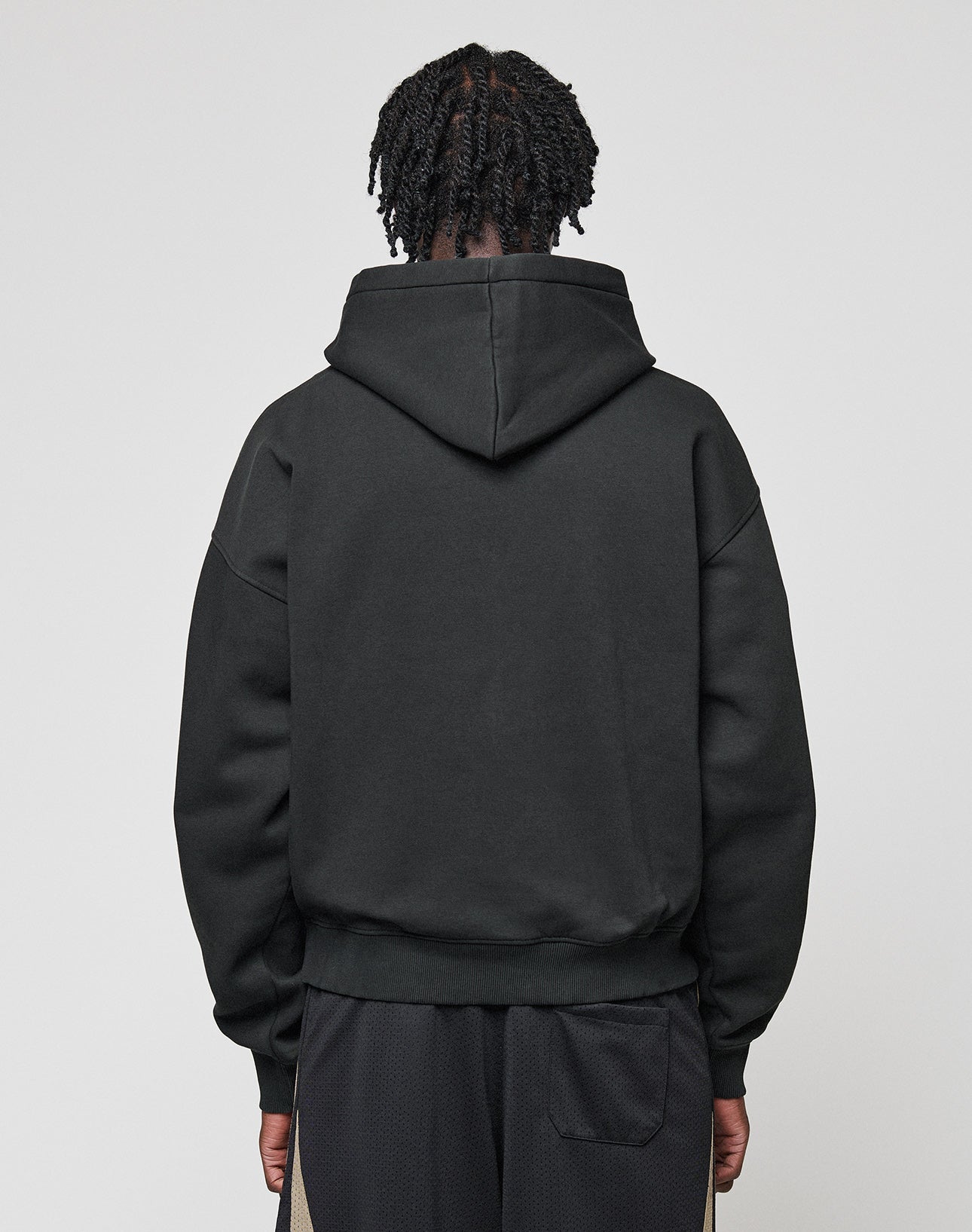 A person with short, twisted hair stands facing away from the camera, wearing the LIVE FAST DIE YOUNG Basic Blank Hooded sweatshirt in black cotton and black pants with a subtle Flag-Label detail, set against a plain light gray background.