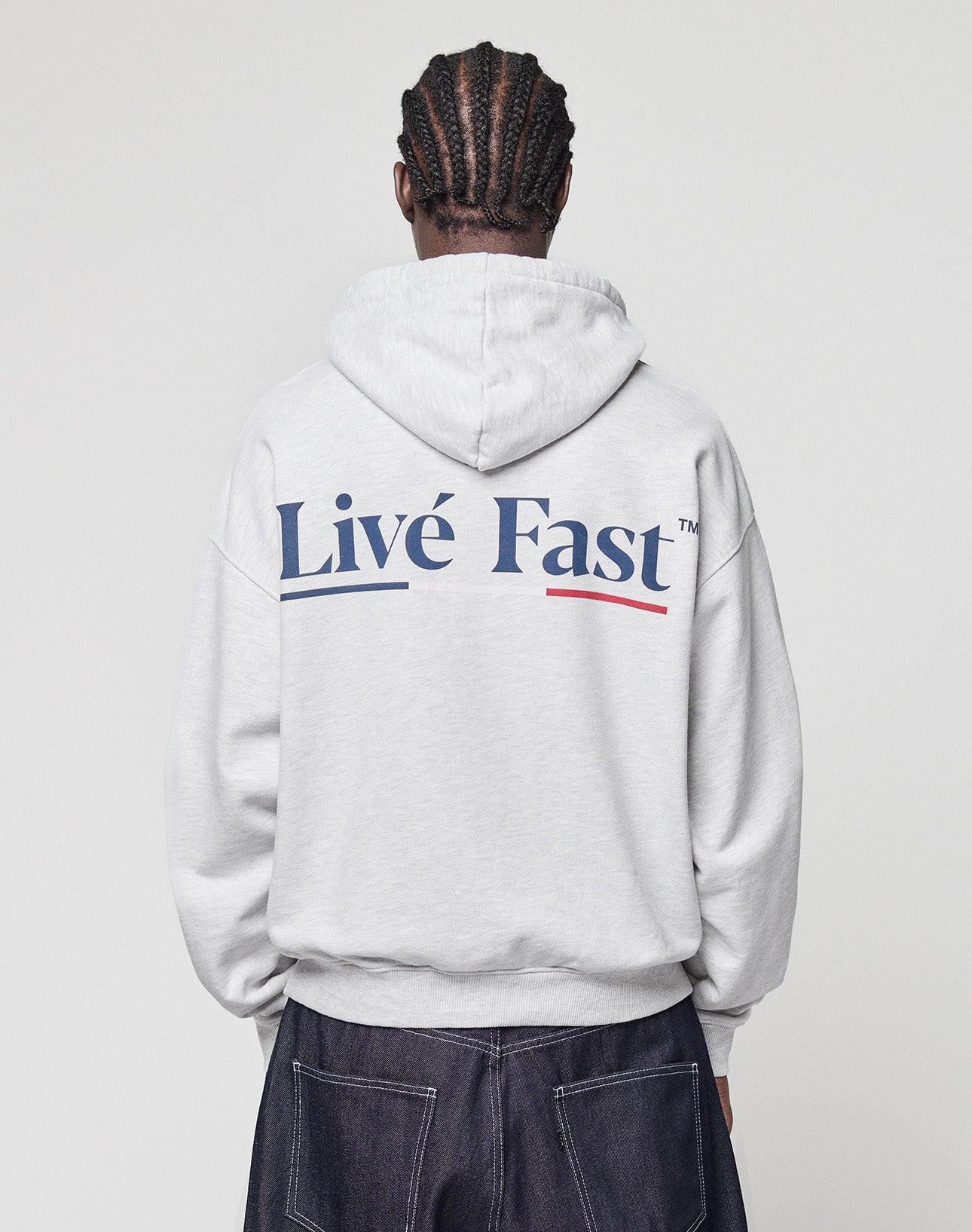 A person with braided hair, seen from behind, wears a light gray LIVE FAST DIE YOUNG Live Fast Paris Hooded pullover with "Livé Fast" in bold blue letters on the back and dark pants with visible stitching.