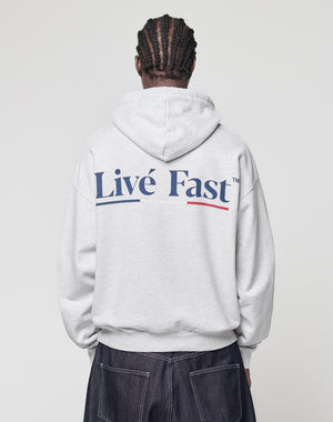 A person with braided hair, seen from behind, wears a light gray LIVE FAST DIE YOUNG Live Fast Paris Hooded pullover with "Livé Fast" in bold blue letters on the back and dark pants with visible stitching.