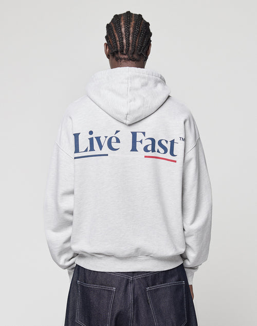 A person with braided hair, seen from behind, wears a light gray LIVE FAST DIE YOUNG Live Fast Paris Hooded pullover with "Livé Fast" in bold blue letters on the back and dark pants with visible stitching.