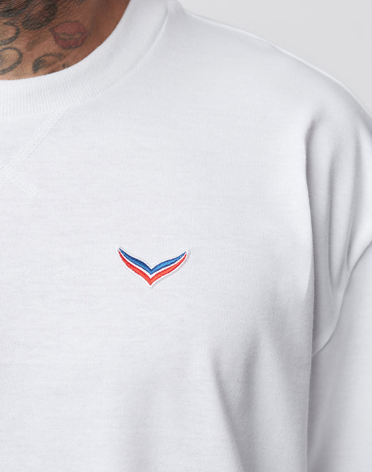 Close-up of a person wearing the LFDY x Trigema Tennis Tee by LIVE FAST DIE YOUNG, an oversize fit white sweatshirt featuring a small red, blue, and black V-shaped logo on the chest. The person’s neck tattoos are partly visible. Limitierte Auflage.