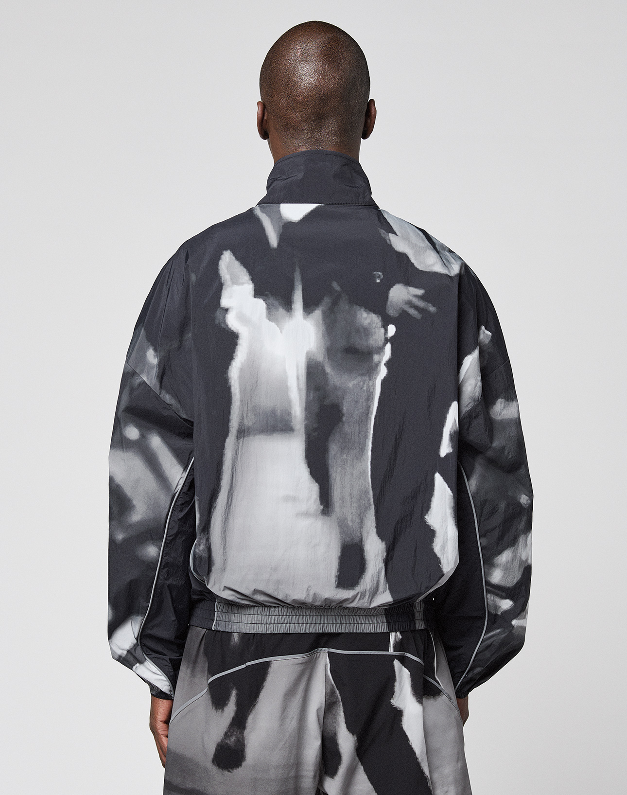 A person with closely shaved hair is seen from behind wearing a loose-fitting black and white LFDY Performance Jacket by LIVE FAST DIE YOUNG, along with matching pants featuring a reflective logo print, set against a plain gray background.