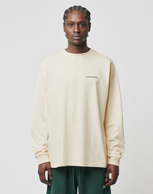 A man with braided hair wears the LIVE FAST DIE YOUNG Basic Longsleeve in cream with a small black logo print on the chest and dark green pants, standing against a plain white background.