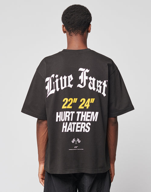 A person with short curly hair is seen from the back wearing the LIVE FAST DIE YOUNG LFDY Haters Tee, a black Oversize Fit T-shirt with bold white and yellow “Live Fast,” “22 24,” and “Hurt Them Haters” prints on front and back.