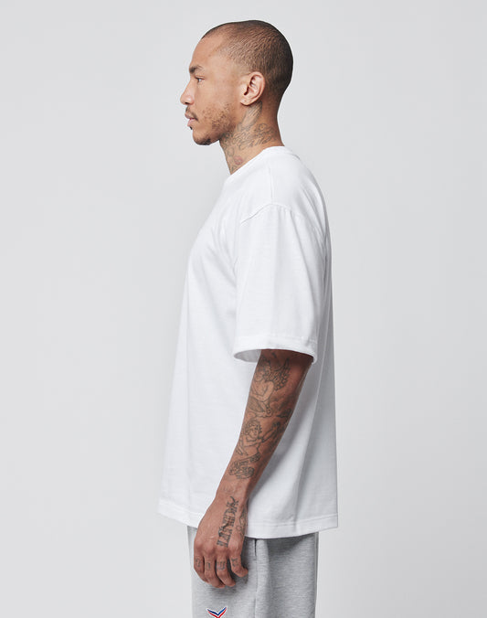 A tattooed man stands in profile facing left, wearing a LIVE FAST DIE YOUNG LFDY x Trigema Tennis Tee and light gray sweatpants against a plain light gray background.