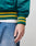 A person sports the LIVE FAST DIE YOUNG Rhine Area College Jacket—green satin with yellow striped cuffs and seitliche Eingrifftaschen—over a white shirt and blue jeans, with focus on their lower arm and hand.