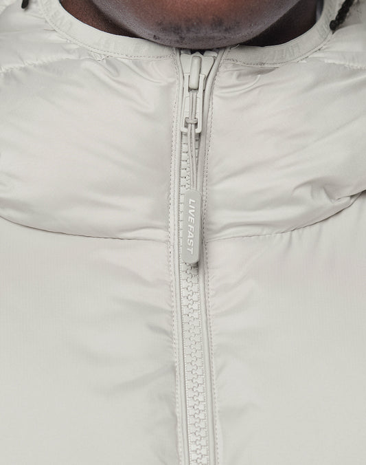Close-up of the light gray Live Fast Puffer Jacket by LIVE FAST DIE YOUNG, featuring a wasserabweisend design and a zipper pull engraved with "LIVE FAST." The lower part of a person's face and neck are partially visible.