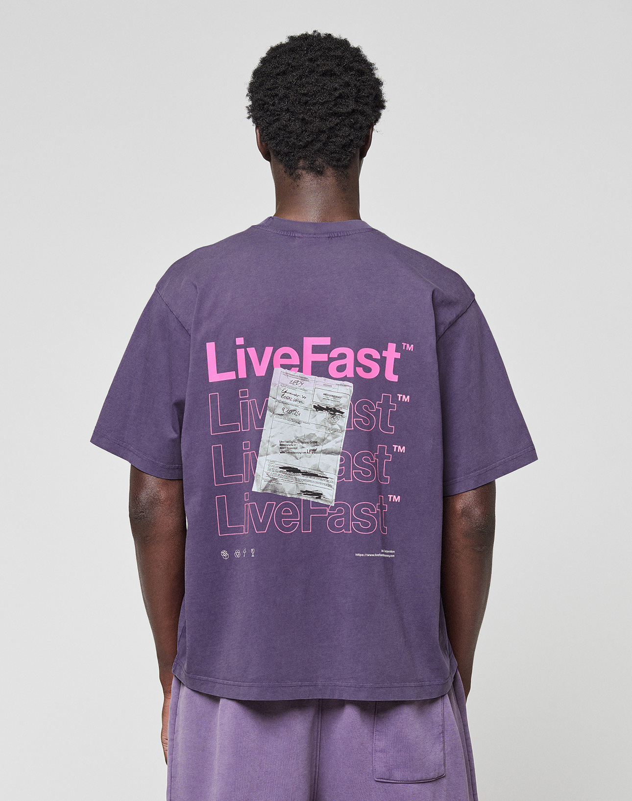A person in a purple Basic Evidence Tee by LIVE FAST DIE YOUNG, made from 100% Baumwolle, featuring repeated “LiveFast” text and a graphic on the back. The front and back print design is shown from behind against a plain background.