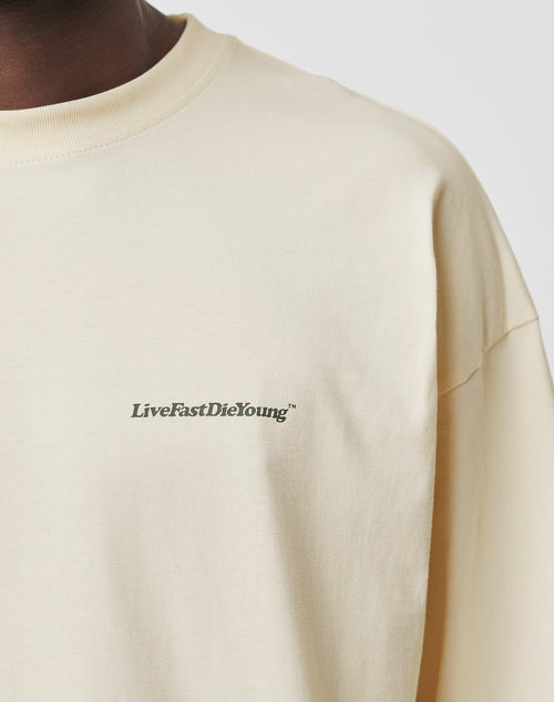 A close-up of a person wearing the LIVE FAST DIE YOUNG Basic Longsleeve in light beige, featuring an oversize fit and a subtle logo-print LiveFastDieYoung™ in dark green on the left chest, made from 100% Baumwolle.