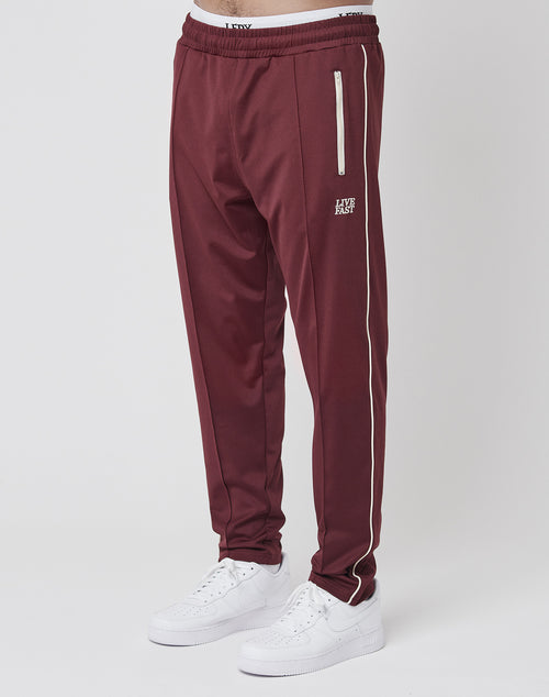 Someone in a regular fit maroon LIVE FAST DIE YOUNG Live Fast Trackjogger with white side piping and zippers, paired with white sneakers, stands against a plain light background.