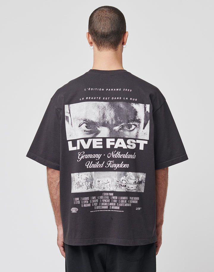 Someone with short hair, facing away, wears the LIVE FAST DIE YOUNG LFDY Cinema Tee—an oversized black Baumwolle t-shirt with a bold backprint of intense eyes, “LIVE FAST,” and the countries Germany, Netherlands, and United Kingdom.