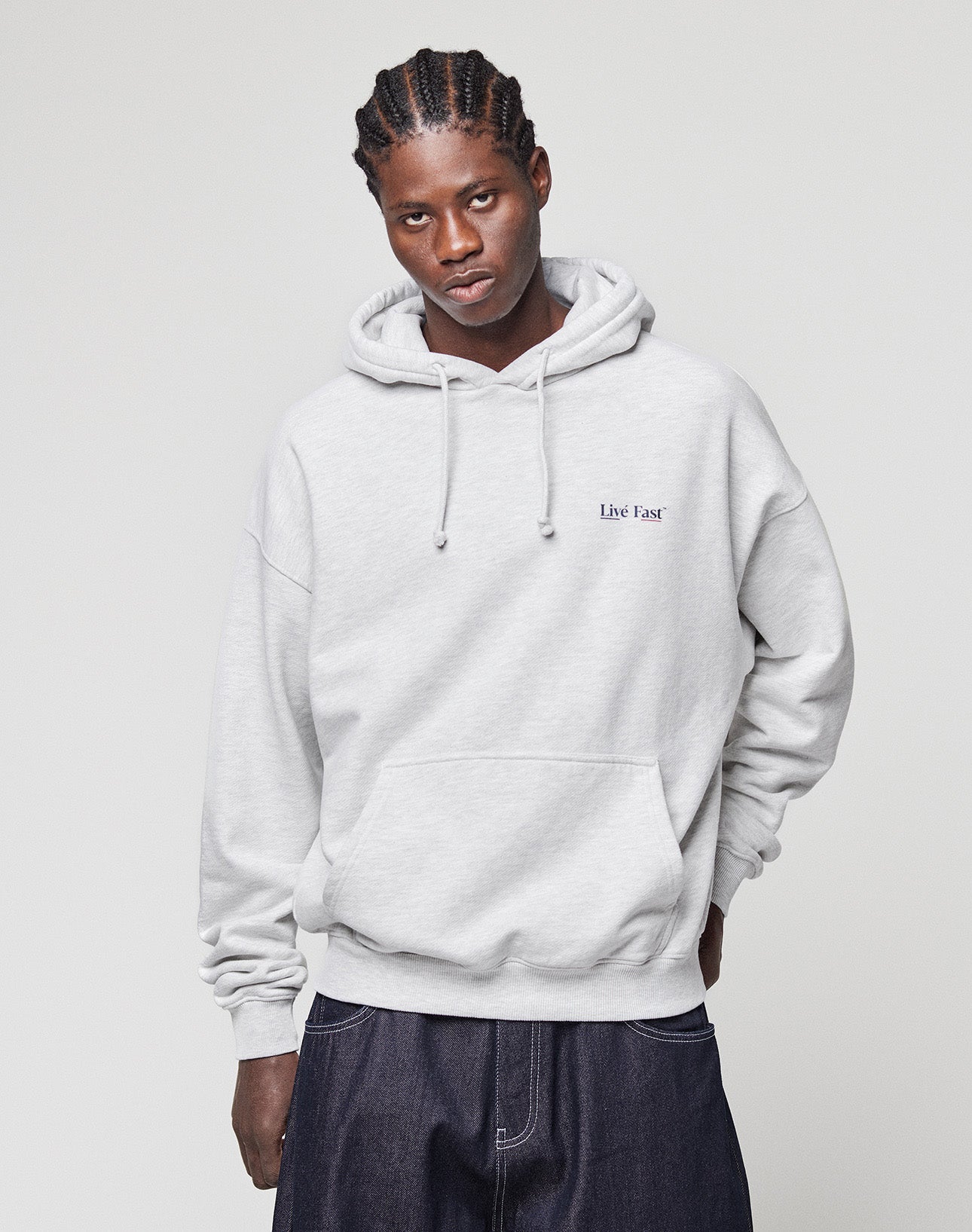 A person in an oversized LIVE FAST DIE YOUNG Live Fast Paris Hooded with front and back print stands against a plain light background. They have braided hair and wear dark, loose-fitting pants.