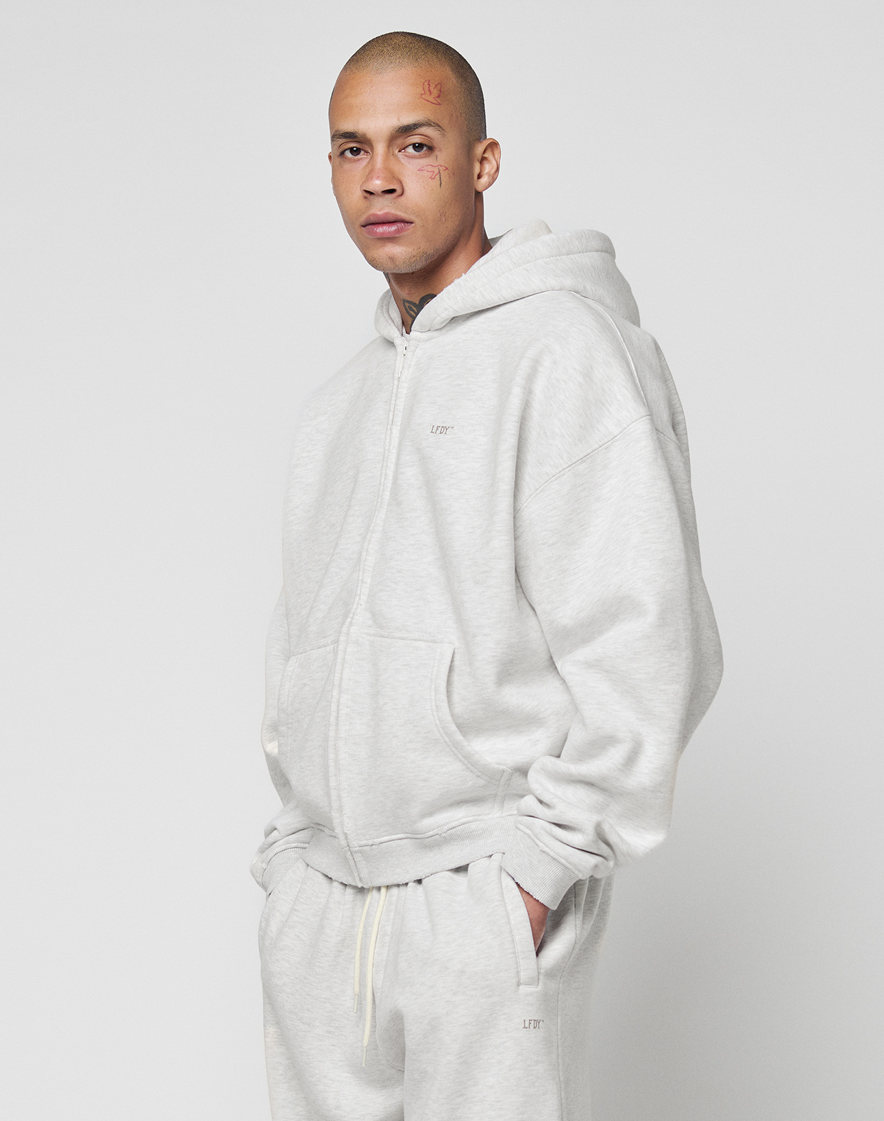 A person wears the LIVE FAST DIE YOUNG Basic Hooded Zip and matching sweatpants, featuring brand logos, standing against a white background with hands in pockets, looking at the camera.
