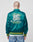 A person with a shaved head wears the LIVE FAST DIE YOUNG Rhine Area College Jacket, a shiny teal nylon bomber with yellow and white building embroidery and LIVE FAST DIE YOUNG RHINE AREA WESTPHALIA on the back, paired with jeans.
