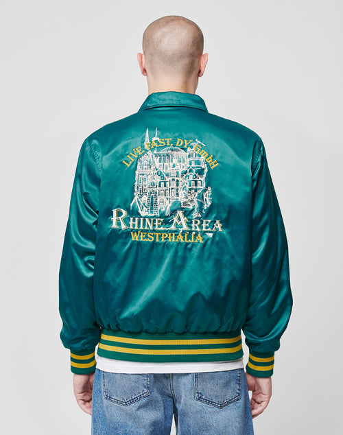 A person with a shaved head wears the LIVE FAST DIE YOUNG Rhine Area College Jacket, a shiny teal nylon bomber with yellow and white building embroidery and LIVE FAST DIE YOUNG RHINE AREA WESTPHALIA on the back, paired with jeans.