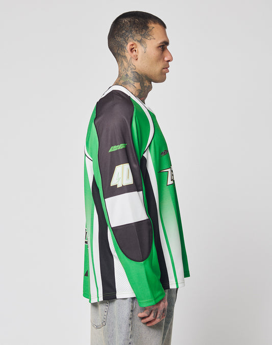 A man with short hair and neck tattoos stands in profile, wearing the LIVE FAST DIE YOUNG LFDY Motorcross Jersey—an oversize green, black, and white long-sleeve with multi-logo prints and 40 on the sleeve—paired with light-washed jeans.